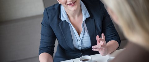 Frau mit roter Kurzhaarfrisur spricht, am Tisch mit Kaffeetasse und Kugelschreiber über Notizen sitzend, zu einer zweiten Frau Frau mit roter Kurzhaarfrisur spricht, am Tisch mit Kaffeetasse und Kugelschreiber über Notizen sitzend, zu einer zweiten Frau